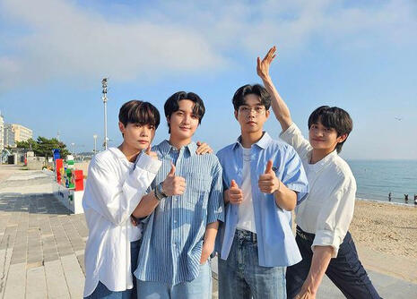 foto de yechan, gwangil, sangyeop y wonsang de la banda coreana lucy en un paseo de una playa. los tres primeros están posando con los pulgares hacia arriba y wonsang está levantando hacia atrás su brazo derecho haciendo una pose más cómica